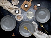 Draufsicht auf stilvolles Tisch-Arrangement mit drei grauen Keramikschalen, goldenem Löffel und Gabel, Teelichtern, Holzbrett und Leinentüchern auf dunkler Oberfläche