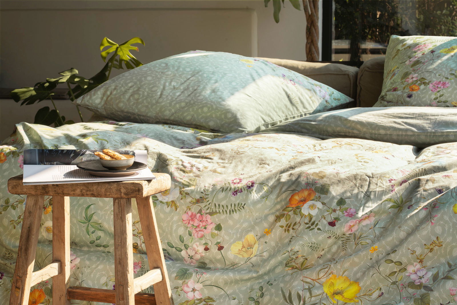 Bettwäsche-Set in sanften Grüntönen mit floralen Mustern, auf einem Bett arrangiert, mit einem Holzhocker im Vordergrund. Perspektive von der Seite.