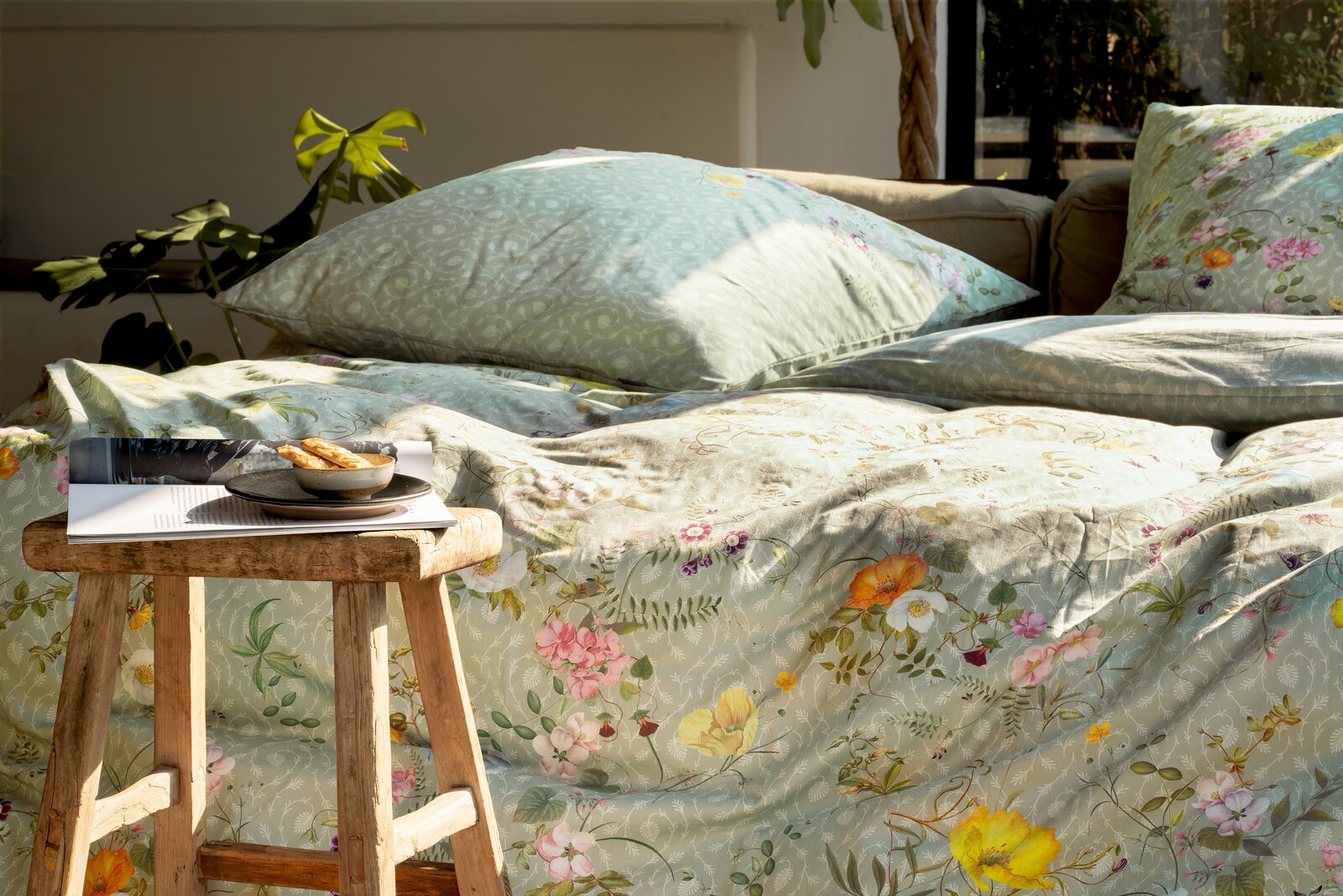 Bettwäsche-Set in sanften Grüntönen mit floralen Mustern, auf einem Bett arrangiert, mit einem Holzhocker im Vordergrund. Perspektive von der Seite.
