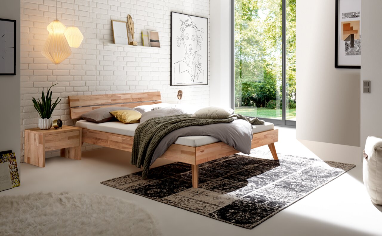 Modernes Schlafzimmer mit einem Bett aus Holz und Metall, seitliche Perspektive. Das Bett hat ein schlichtes Kopfteil, flankiert von einem Nachttisch. Der Raum ist hell und minimalistisch eingerichtet, mit großen Fenstern, die einen Blick ins Grüne bieten