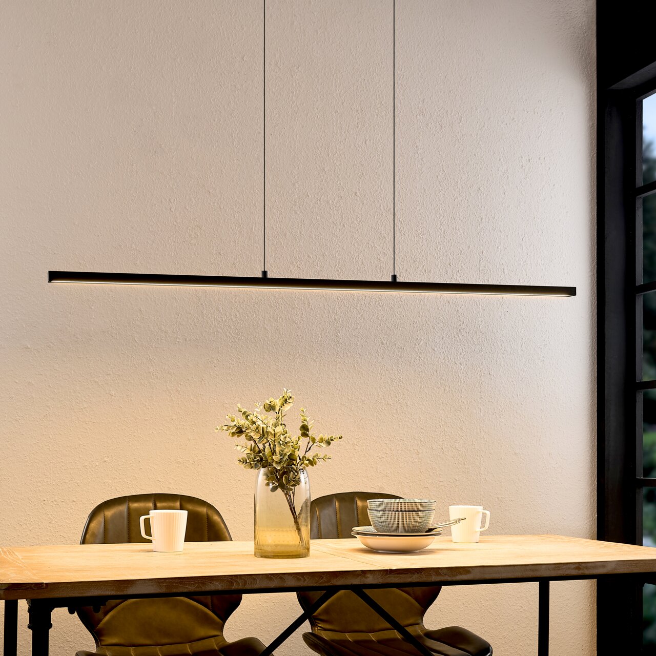 Moderne, slanke LED-hanglamp in zwart als lange lichtbalk boven een houten tafel met decoratie en serviesgoed, van voren gefotografeerd vanuit een licht verhoogd perspectief.