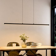 Moderne, slanke LED-hanglamp in zwart als lange lichtbalk boven een houten tafel met decoratie en serviesgoed, van voren gefotografeerd vanuit een licht verhoogd perspectief.