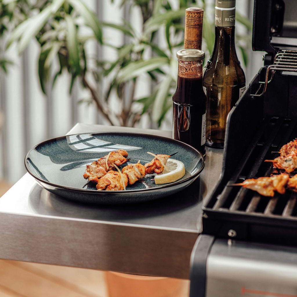 Grauer Speiseteller Lave 28cm mit gegrillten Fleischspießen und Zitronenscheibe, seitliche Perspektive neben einem Grill.