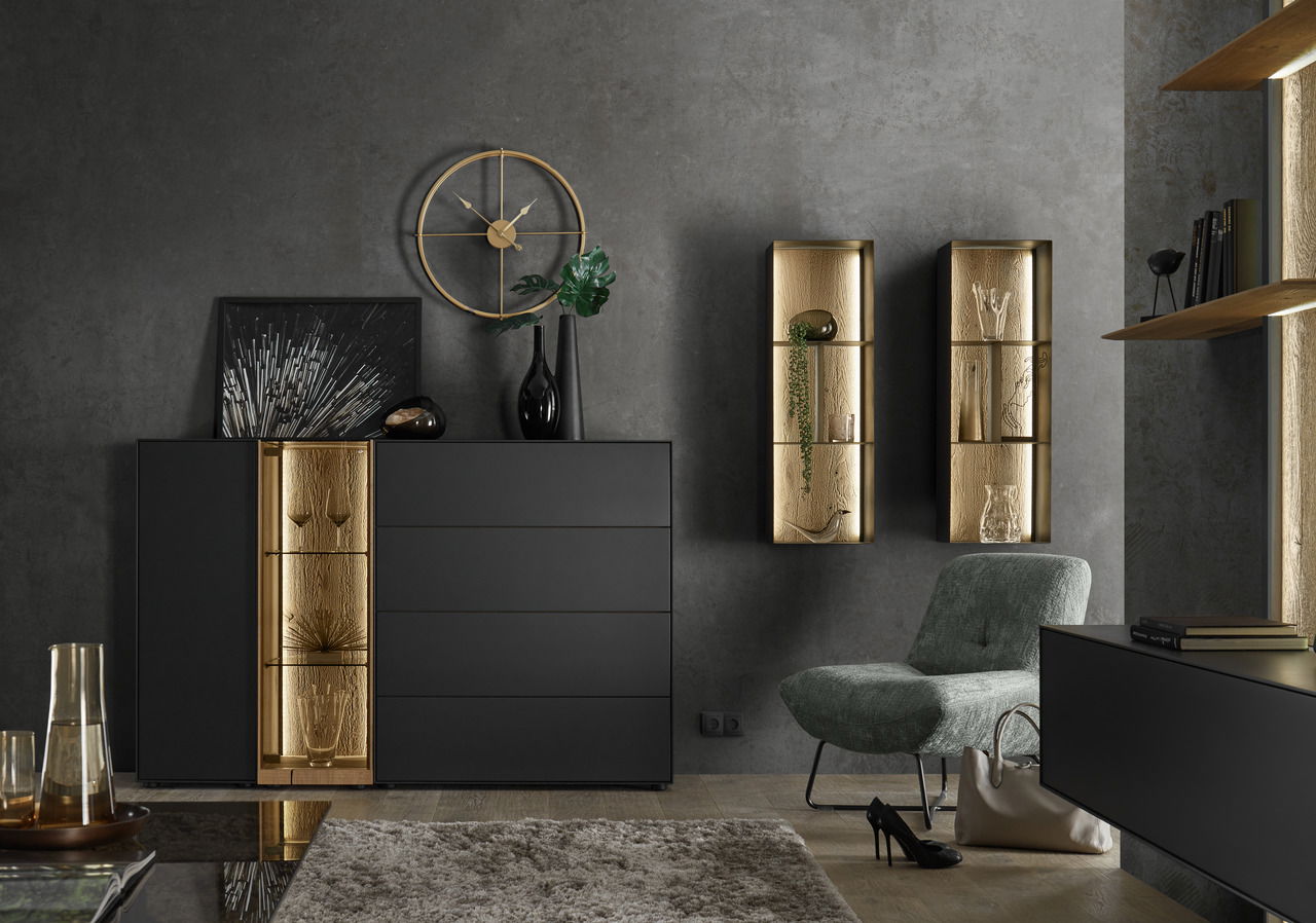 Modernes Highboard in schwarzem Design mit goldenen Akzenten, frontal fotografiert. Dekoriert mit Vasen und Kunstobjekten, umgeben von stilvollen Wandregalen und einem Sessel.