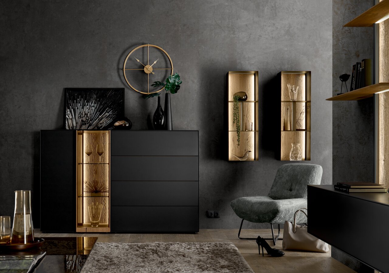 Musterring Hoog dressoir TREVIO Modern hoogkast in zwart met gouden accenten, gefotografeerd vanaf de voorkant. Versierd met vazen en kunstvoorwerpen, omringd door stijlvolle wandplanken en een fauteuil.