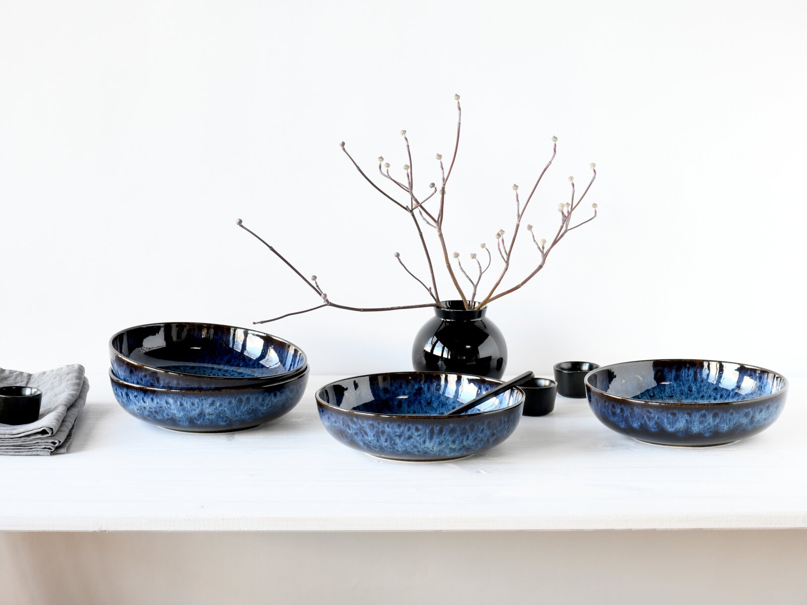 Blau glasierte Keramik-Schalen im Set auf weißem Tisch, dekoriert mit schwarzer Vase und Zweigen, Frontansicht auf Augenhöhe