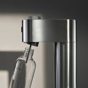 Nahaufnahme eines Wassersprudlers aus Edelstahl mit einer angeschlossenen Glasflasche, seitliche Perspektive.