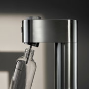 Nahaufnahme eines Wassersprudlers aus Edelstahl mit einer angeschlossenen Glasflasche, seitliche Perspektive.