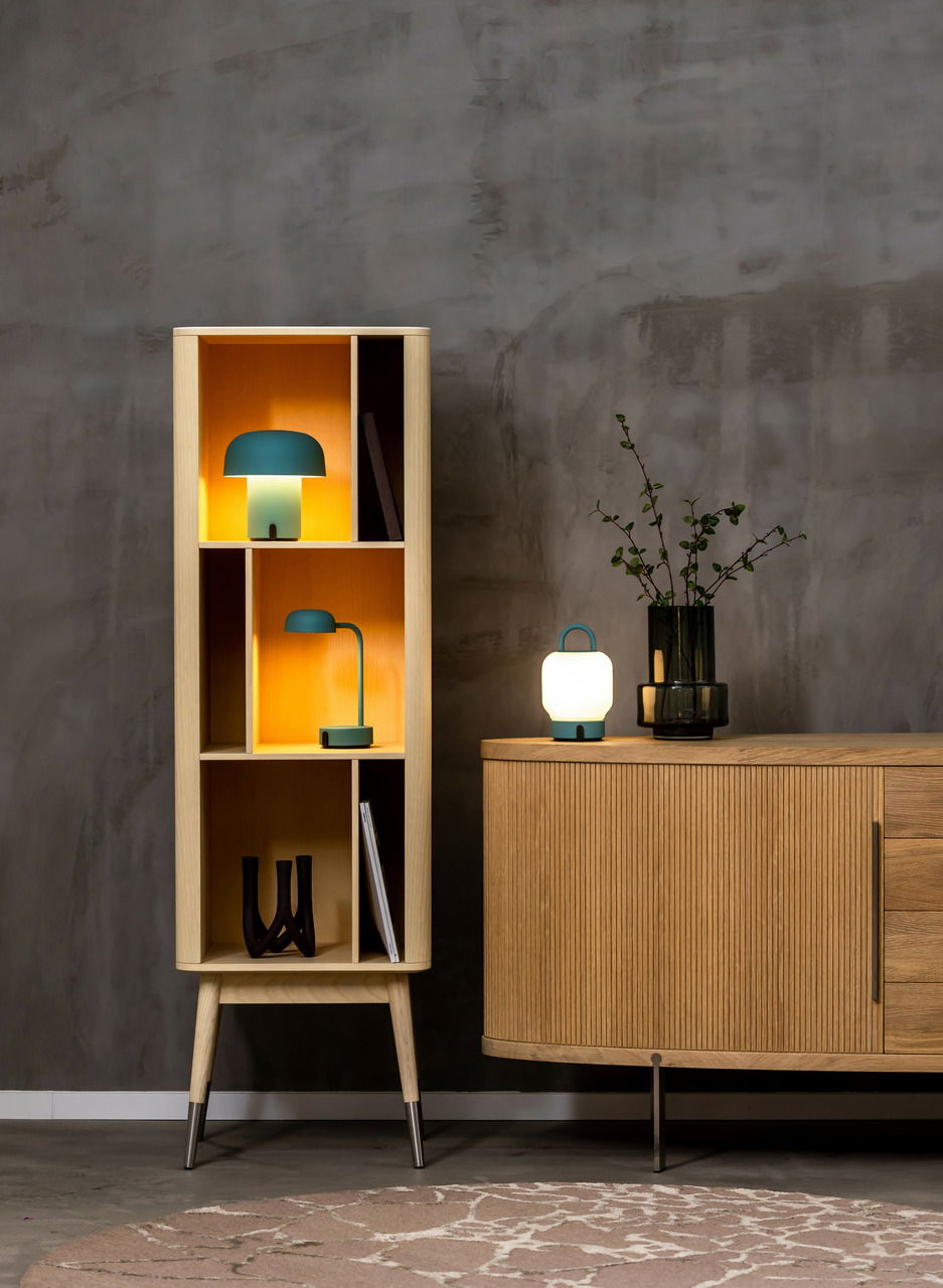 Ein modernes Wohnzimmer mit einem hohen, offenen Regal aus hellem Holz, das zwei stilvolle Lampen in Smokey Teal zeigt. Eine Tischlampe und eine Pilzlampe leuchten warm. Rechts steht ein Sideboard mit einer weiteren Lampe und einer Vase. Perspektive: Fron