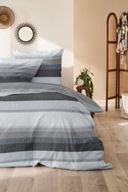 Bettwäsche-Set in Grau- und Blautönen mit Streifenmuster, auf einem Bett in einem stilvoll eingerichteten Raum mit Holzboden und Dekorationen, aus der Perspektive von vorne fotografiert.