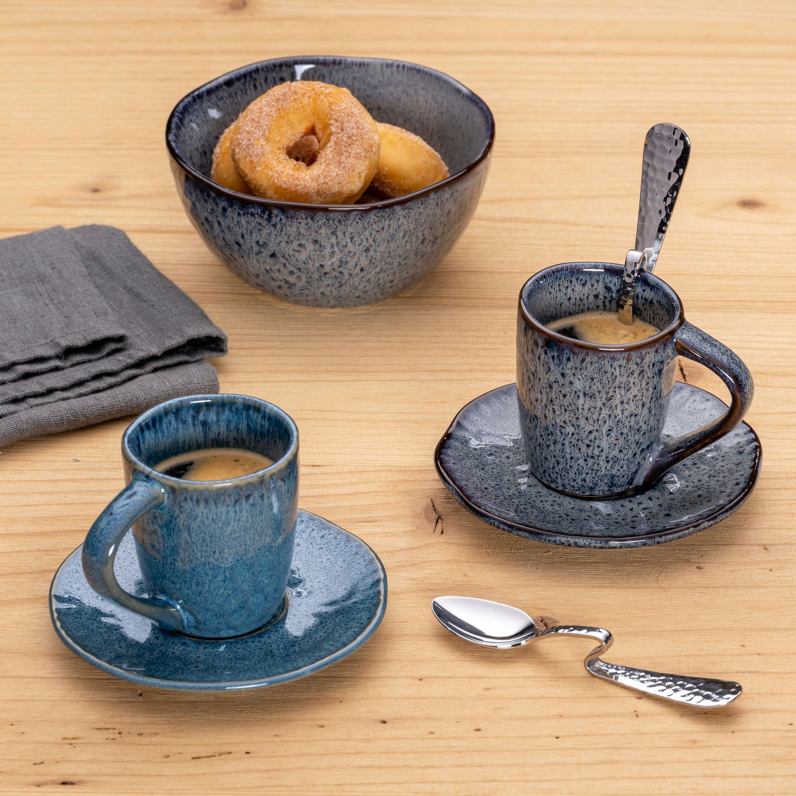 Leonardo Espressolöffel Gebogener Espressolöffel aus Edelstahl auf einem Holztisch, neben zwei blauen Espressotassen und einer Schale mit Donuts, aus der Vogelperspektive.