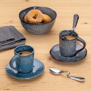 Gebogener Espressolöffel aus Edelstahl auf einem Holztisch, neben zwei blauen Espressotassen und einer Schale mit Donuts, aus der Vogelperspektive.