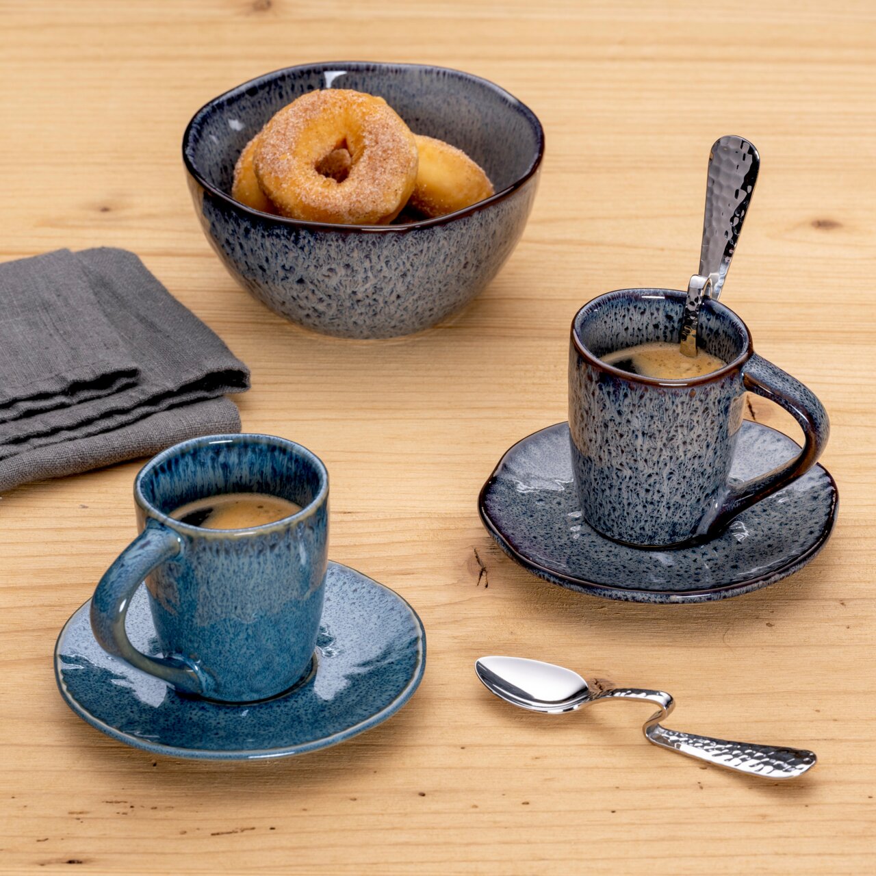 Leonardo Espressolöffel Gebogener Espressolöffel aus Edelstahl auf einem Holztisch, neben zwei blauen Espressotassen und einer Schale mit Donuts, aus der Vogelperspektive.