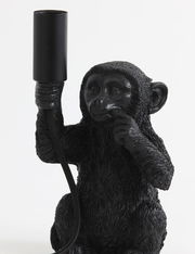 Schwarze Tischleuchte in Form eines sitzenden Affen, der eine Lampenfassung in der Hand hält. Frontalansicht.