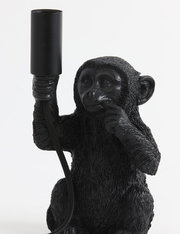 Schwarze Tischleuchte in Form eines sitzenden Affen, der eine Lampenfassung in der Hand hält. Frontalansicht.