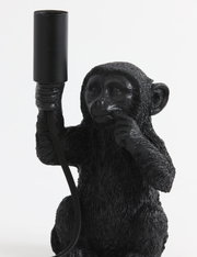 Schwarze Tischleuchte in Form eines sitzenden Affen, der eine Lampenfassung in der Hand hält. Frontalansicht.