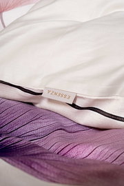 Nahaufnahme aus leicht schräger Seitenperspektive: weißes Kissen mit schwarzer Paspel und ESSENZA-Label auf lila-rosa gemusterter Bettwäsche