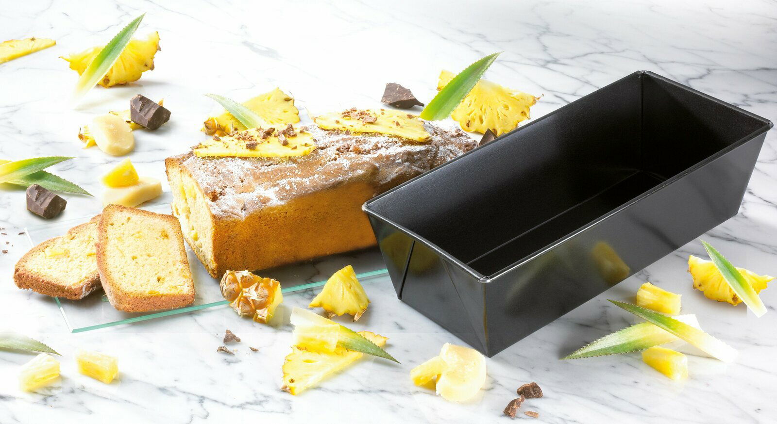 Kastenform 25cm BackFreude aus schwarzem Material, neben einem frisch gebackenen Kuchen mit Ananas und Schokoladenstücken, auf einer Marmoroberfläche. Perspektive von oben.