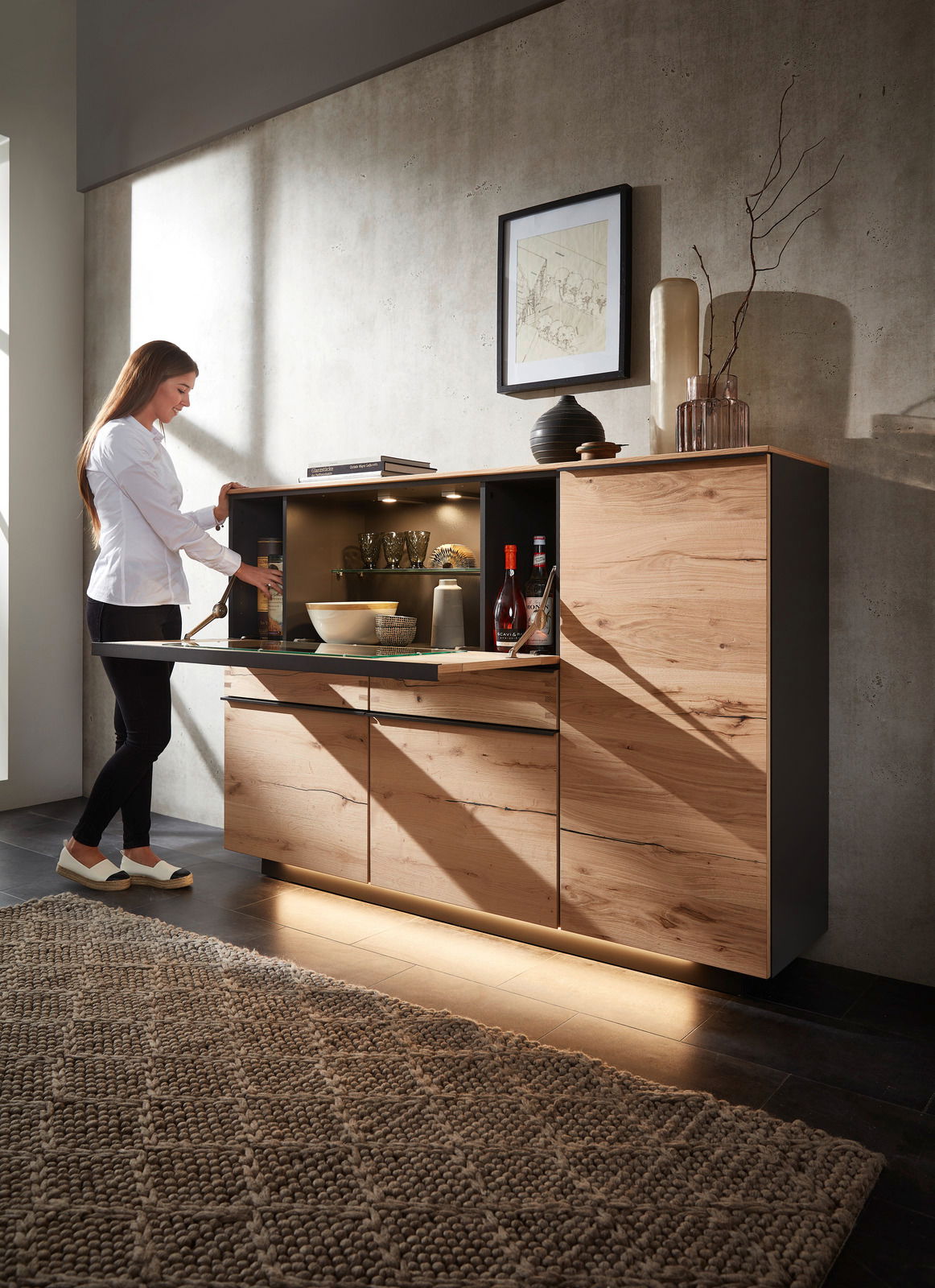 Highboard aus Holz mit geöffneter Klappe, beleuchtetem Innenraum und dekorativen Gegenständen, seitliche Perspektive
