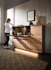 Highboard aus Holz mit geöffneter Klappe, beleuchtetem Innenraum und dekorativen Gegenständen, seitliche Perspektive