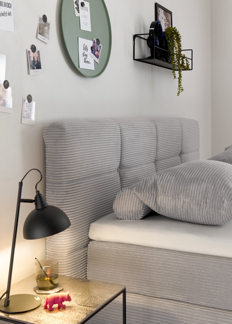 Zijaanzicht van een grijs boxspringbed met geribbelde stoffen bekleding, versierd met kussens. Op de voorgrond een nachtkastje met een lamp en decoratie.