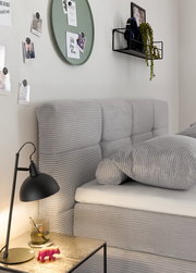 Seitliche Perspektive eines grauen Boxspringbetts mit geripptem Stoffbezug, dekoriert mit Kissen. Im Vordergrund ein Nachttisch mit Lampe und Dekoration.