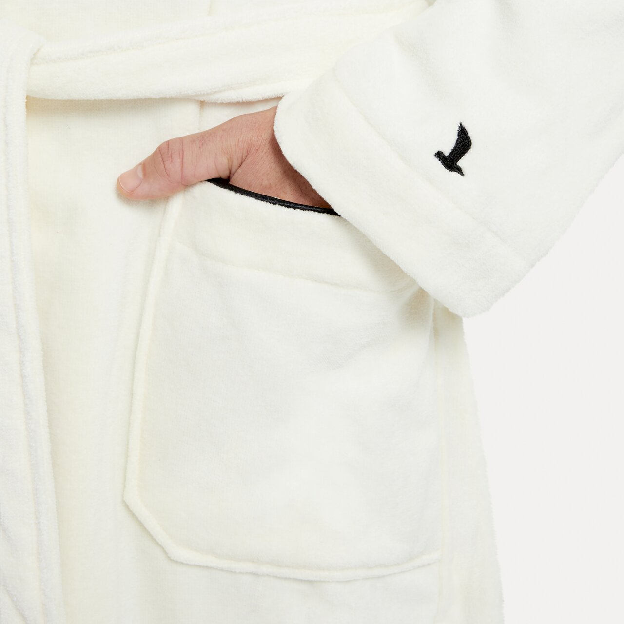 Nahaufnahme eines weißen Bademantels mit Schalkragen in Größe XL, zeigt eine Hand in der Tasche und ein gesticktes Logo auf dem Ärmel.