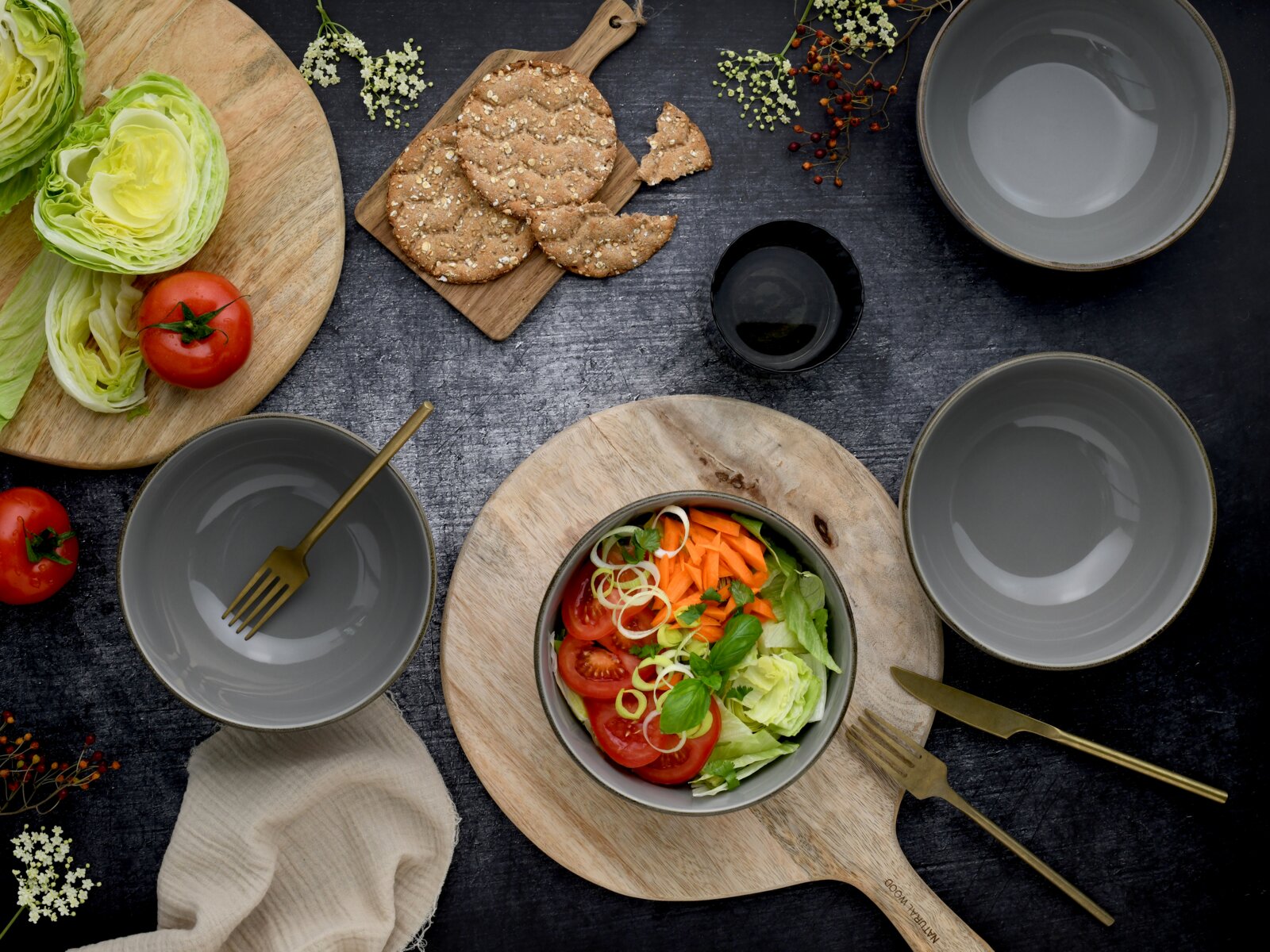 Draufsicht (Flatlay) auf einen gedeckten Tisch mit Schüssel frischem Salat (Tomaten, Karotten, Salat, Kräuter), grauen Schalen, goldfarbenem Besteck, Knäckebrot auf Holzbrett sowie Salatkopf und Tomaten auf Schneidebrett.