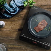 Einzelkochplatte mit Pfanne, in der ein Stück Fleisch und Kräuter braten, aus der Vogelperspektive auf einem Holztisch.