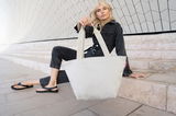 Frau in schwarzem Jeans-Outfit sitzt auf Treppe und hält eine große, sandfarbene Shopper-Tasche mit Fischgrätenmuster. Perspektive von vorne.