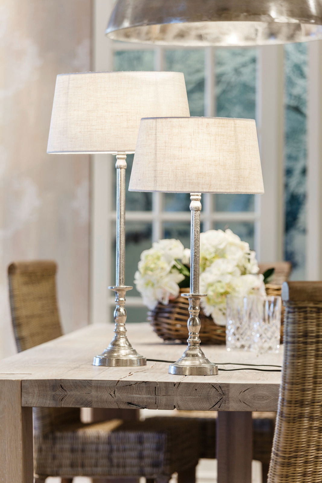 Zwei elegante Tischlampen mit silbernen, runden Sockeln und cremefarbenen, ellipsenförmigen Lampenschirmen auf einem Holztisch in einem gemütlichen Esszimmer. Perspektive von der Seite.