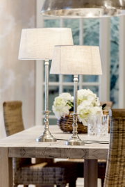 Zwei elegante Tischlampen mit silbernen, runden Sockeln und cremefarbenen, ellipsenförmigen Lampenschirmen auf einem Holztisch in einem gemütlichen Esszimmer. Perspektive von der Seite.