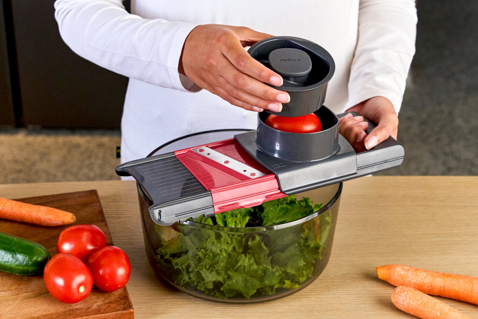 Schräg von vorn fotografierter Gemüsehobel/Mandoline mit Auffangschüssel; Person schneidet eine Tomate über einer Schüssel mit Salat, daneben liegen Gurke, Tomaten und Karotten auf dem Tisch.
