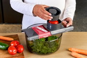 Schräg von vorn fotografierter Gemüsehobel/Mandoline mit Auffangschüssel; Person schneidet eine Tomate über einer Schüssel mit Salat, daneben liegen Gurke, Tomaten und Karotten auf dem Tisch.