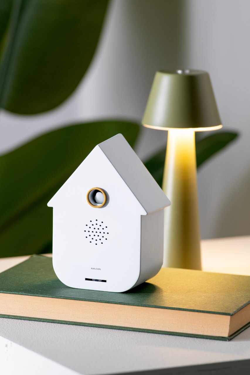 Weiße moderne Soundbox in Form eines Hauses, frontal auf einem Buch platziert, mit einer Lampe im Hintergrund.