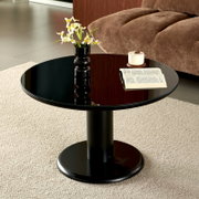 Runder, schwarzer Couchtisch mit glänzender Tischplatte und zentralem Standfuß, dekoriert mit schwarzer Vase mit gelb-weißen Blumen, Tasse und aufgeschlagenem Buch; Perspektive: leicht erhöhte Frontansicht.