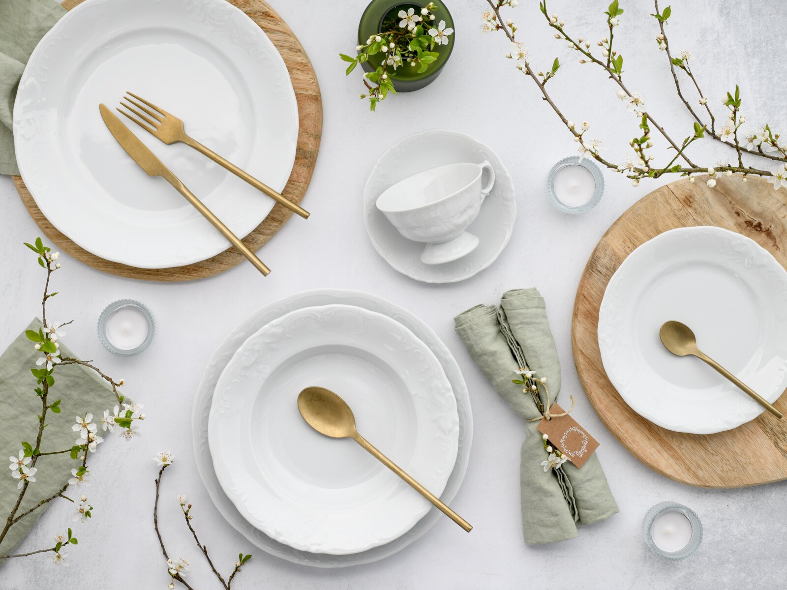 Von oben fotografiertes, elegantes Tischgedeck mit weißen Porzellantellern und -schalen mit Reliefrand, goldfarbenem Besteck, Holzuntersetzern, grünen Leinenservietten, Teelichtern und blühenden Zweigen auf hellem Hintergrund