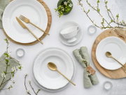 Von oben fotografiertes, elegantes Tischgedeck mit weißen Porzellantellern und -schalen mit Reliefrand, goldfarbenem Besteck, Holzuntersetzern, grünen Leinenservietten, Teelichtern und blühenden Zweigen auf hellem Hintergrund