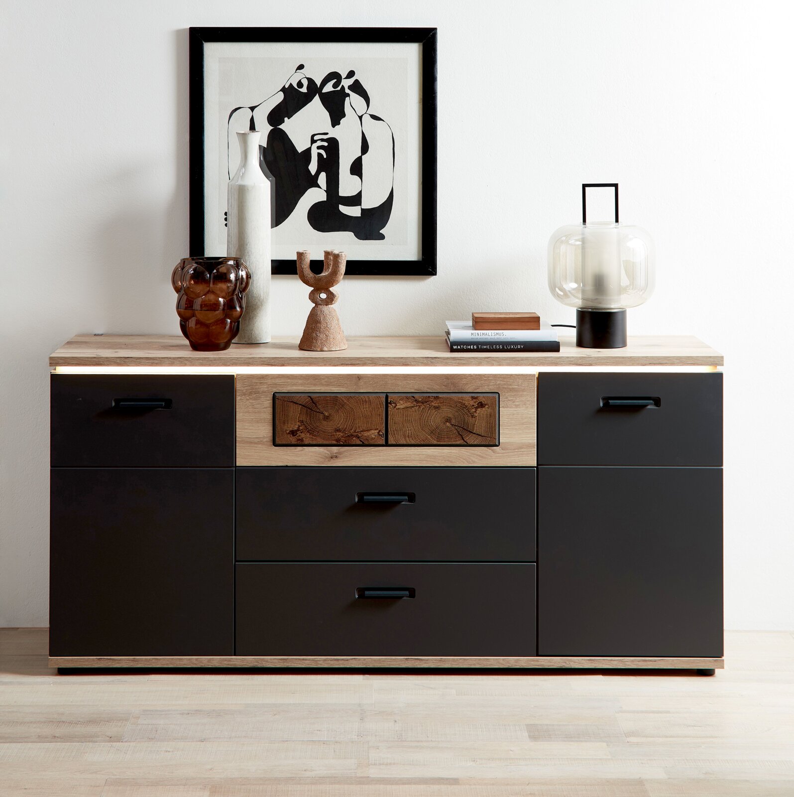 Moderne Sideboard-Kommode in Schwarz mit Holzplatte und mittlerer Holz-Schubladenfront, dekoriert mit Vase, Skulptur, Büchern und Tischlampe; frontale Perspektive auf das Möbel vor weißer Wand.