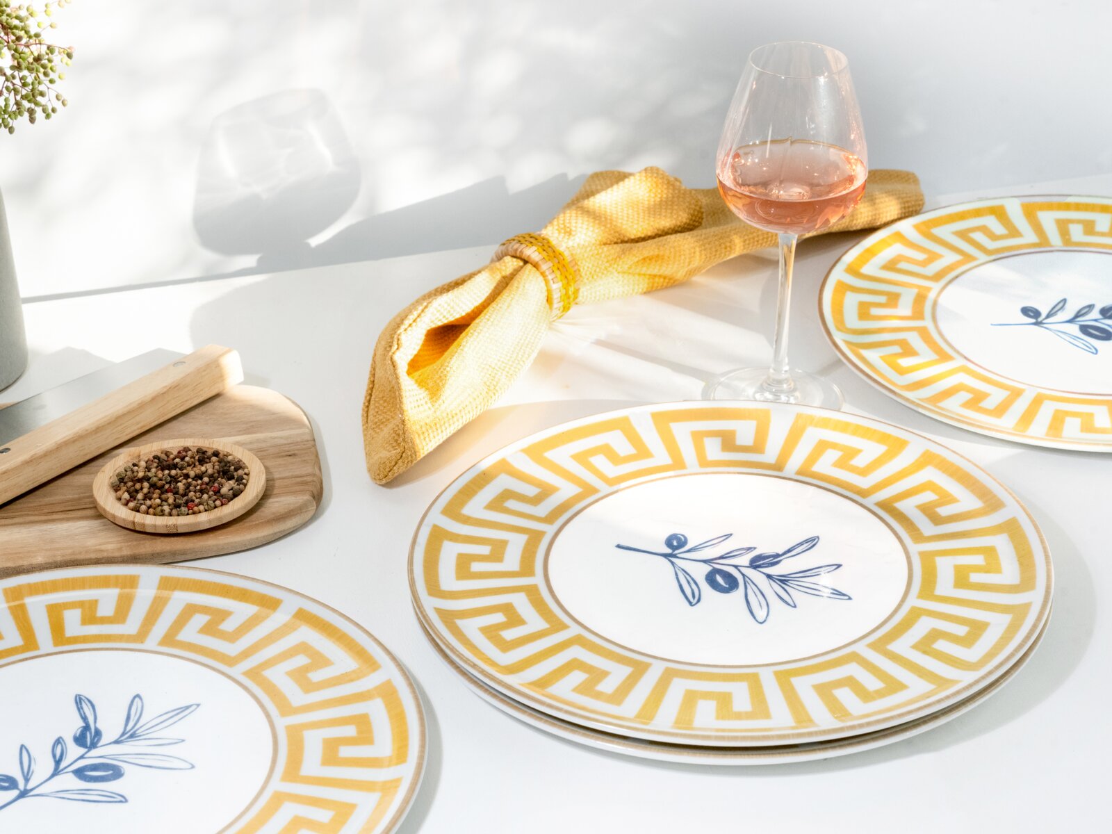 Sommerlich gedeckter Tisch mit weißem Geschirr: Teller mit gelbem Mäander-Rand und blauem Olivenzweig-Motiv, gelbe Stoffserviette mit Ring, Weinglas mit Rosé sowie Holzbrett mit Gewürzen – Perspektive: leicht von oben, schräger Blickwinkel.