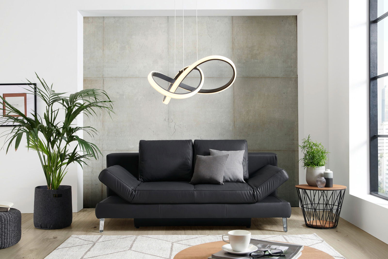 Novel Schlafsofa LAVINIA Schwarzes Querschläfer-Sofa in modernem Wohnzimmer, frontal fotografiert, mit Pflanzen und Couchtisch.