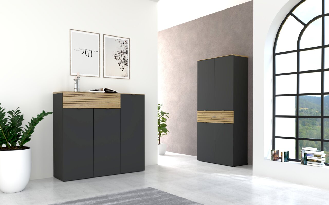 Moderne Garderobenmöbel in Graphit mit Holzdetails, bestehend aus einem niedrigen und einem hohen Schrank, in einem hellen Raum mit großen Fenstern, Pflanzen und Wanddekorationen. Perspektive von vorne links.