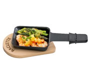 Raclette-Pfännchen mit Griff, gefüllt mit Garnelen, Spargel und geschmolzenem Käse, auf einem ovalen Holzbrettchen mit der Aufschrift 'Raclette', aus der Vogelperspektive.