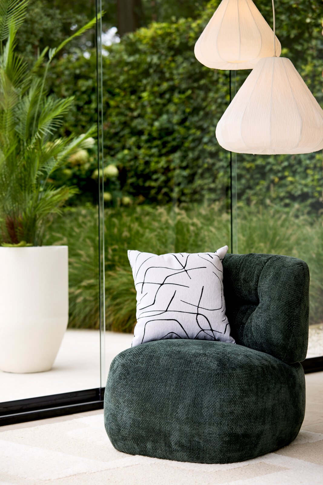 Donkergroene, organisch gevormde loungestoel van voren in driekwartperspectief, gestyled met een wit sierkussen in een lichte woonruimte met zicht op de tuin.