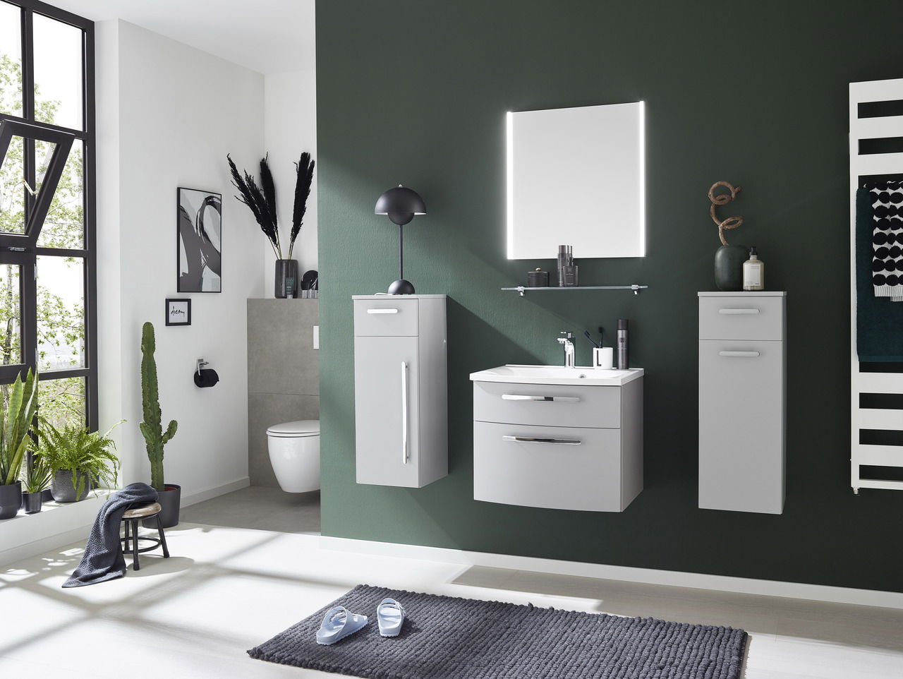 Moderner Badblock 3-teilig in grauer Farbe, bestehend aus einem Waschbeckenunterschrank, einem Hängeschrank und einem Hochschrank, vor einer dunkelgrünen Wand. Perspektive: Frontalansicht.