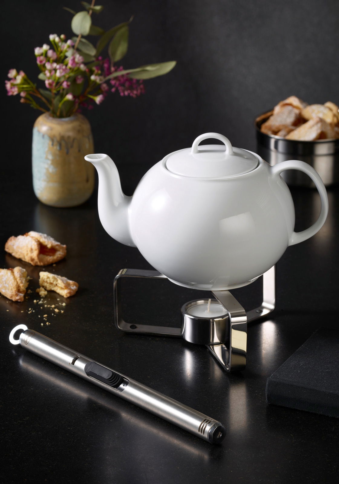 Weißes Teekännchen auf einem modernen Stövchen mit Teelicht, seitliche Perspektive, umgeben von Gebäck, Blumen und einem Feuerzeug.