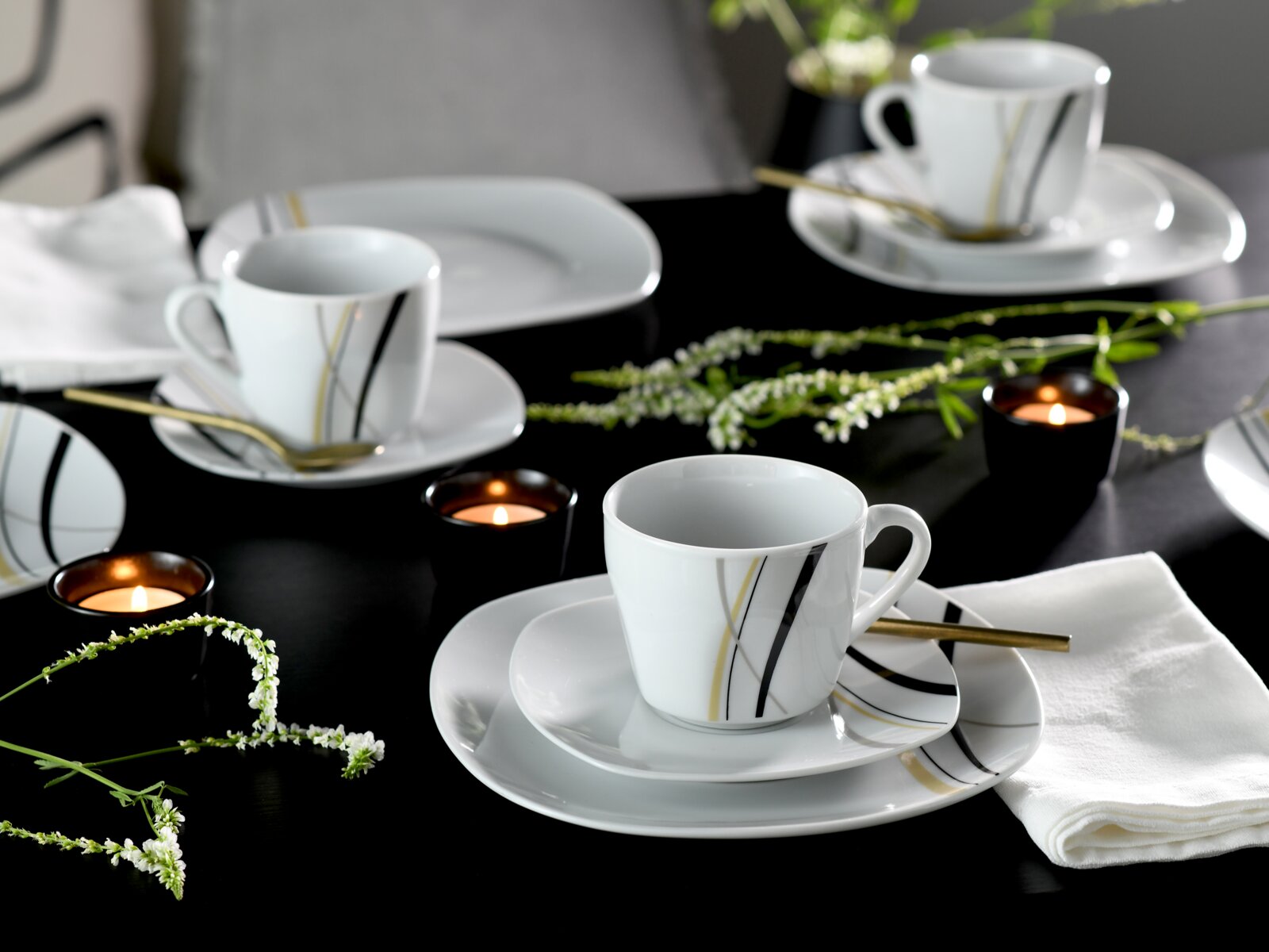 Modernes weißes Porzellan-Kaffeeservice mit Tassen, Untertassen und Tellern mit schwarzen und goldenen Linien, dekoriert mit Kerzen und Blumen auf dunklem Tisch, Perspektive: leicht erhöhte schräge Ansicht von vorne