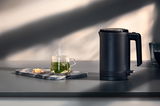 Schwarzer Wasserkocher mit 0,8L Fassungsvermögen, seitliche Perspektive, neben einer Tasse dampfendem Tee auf einem Tisch.