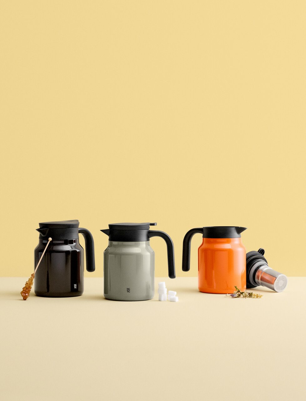 Drie THERM-IT-thermoskannen in verschillende kleuren (zwart, grijs, oranje) op een tafel tegen een gele achtergrond, van voren gefotografeerd.