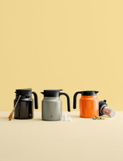 Drie THERM-IT-thermoskannen in verschillende kleuren (zwart, grijs, oranje) op een tafel tegen een gele achtergrond, van voren gefotografeerd.